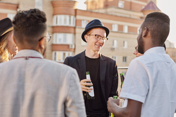 Multiethnic group of young stylish men and women standing on rooftop terrace, drinking beer and having friendly conversation