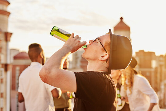 Young ginger man in bowler hat and glasses drinking beer from bottle while hanging out with friends on rooftop terrace on sunny evening