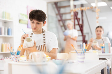 Skilled interested young artist sitting at table in pottery workshop, painting designs on ceramic cup. Art craft activity concept..