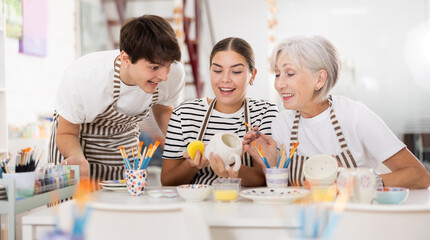 Friendly older mother and young adult daughter and son, wearing aprons, talking cheerfully while working on painting ceramic dishware in family pottery workshop. Generational collaboration concept..