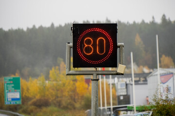 Glowing red digital road sign displaying 80 km/h limit against autumn landscape background showing modern traffic control technology and organized driving system in contemporary infrastructure