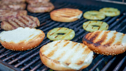Grilled burger patties and toasted sesame buns with pineapple slices on barbecue grill, showcasing summer cooking and outdoor dining experience