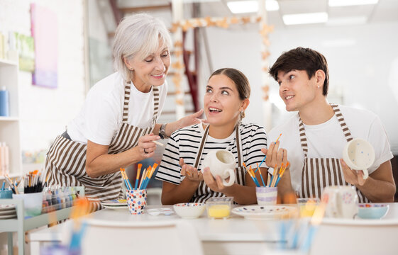 Enthusiastic senior female teacher sharing experience pottery craftsmanship, assisting happy young guy and girl, painting ceramic mugs and plates at table in workshop