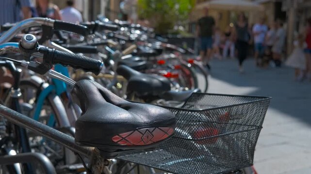 Close-up on bike saddle with people walking in the background. Bike saddles and handlebars positioned in sharp focus with pedestrians blurring behind, capturing urban cycling culture street scene