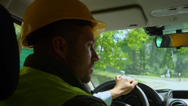 Rear of man worker in yellow helmet and uniform driving a car at a construction site. Close up of male builder in hard hat in vehicle going at workplace. Work trip, constructor job