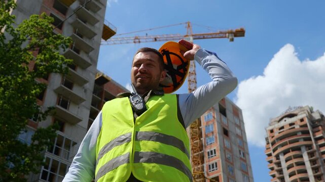 Bottom view of happy handsome young Caucasian man builder standing outdoor taking off yellow helmet on sunny day. Close up. Construction business concept, architect industry
