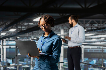 Black female programmer working on laptop in data center, handling maintenance tasks to ensure...