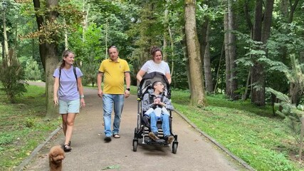 Family with a boy with a central nervous system disorder walking in the park. Family with a boy with inclusion. A boy with a cerebral palsy in a wheelchair walks with his family in the park