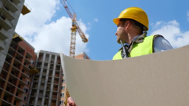 Handsome young male constructor engineer wearing helmet and uniform holding in hands work project and looking at construction site. Bottom view. Architecture inspector work