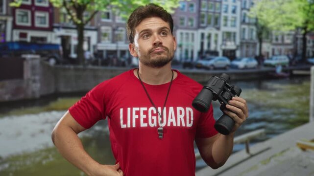 Young hispanic man lifeguard holding binoculars and whistle, showing thumb down gesture on street by a canal with buildings behind him; safety alert.