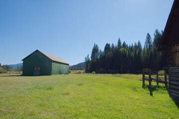 Obraz premium Rustic Wooden Barn With Weathered Logs On a Bright Green Field Under Clear Blue Sky. Historic Rustic Ant Flat Kootenai National Forest in Montana. Evoking countryside charm and rural life in a serene