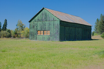 Obraz premium Rustic Wooden Barn With Weathered Logs On a Bright Green Field Under Clear Blue Sky. Historic Rustic Ant Flat Kootenai National Forest in Montana. Evoking countryside charm and rural life in a serene