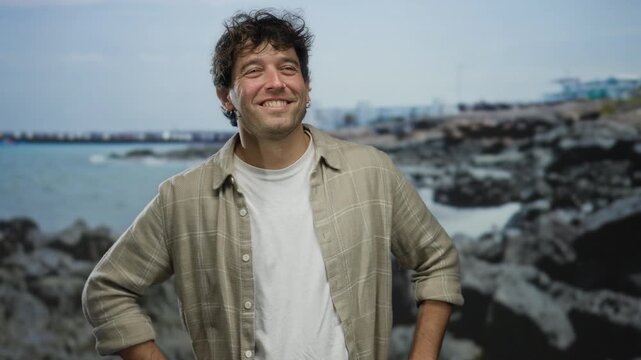 Smiling man with arms crossed stands confidently on a rocky beach with the sea in the background, exuding warmth and charm in a relaxed coastal setting.