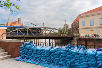 Blue sandbags flood barrier on riverside embankment with historic cathedral, metal bridge and tram in background on cloudy day. Concept of urban flood protection and emergency disaster prevention.