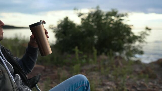 A man is resting on a car trip on the coast of a lake, ocean or sea. He has a seat in a folding chair. He is holding a thermos or thermo mug