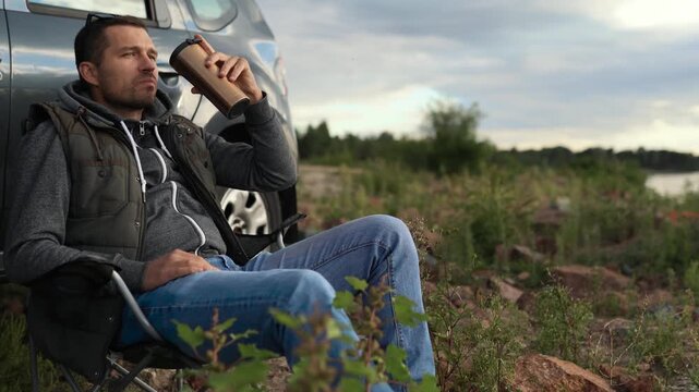A man is resting on a car trip on the coast of a lake, ocean or sea. He has a seat in a folding chair. He is holding a thermos or thermo mug