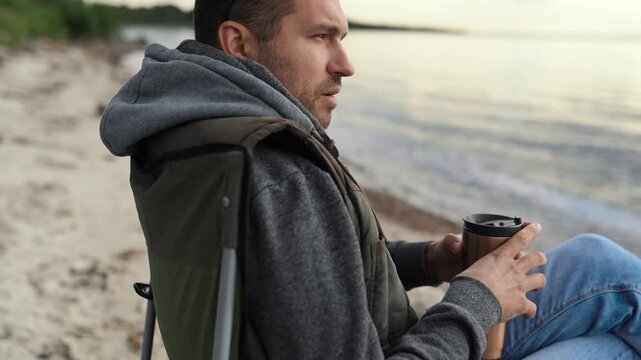 A man is resting on a car trip on the coast of a lake, ocean or sea. He has a seat in a folding chair.