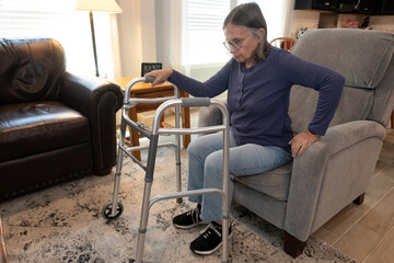 Elderly Woman Holds Onto Her Walker With One Hand While She Gets up Out of a Chair in Her Living Room