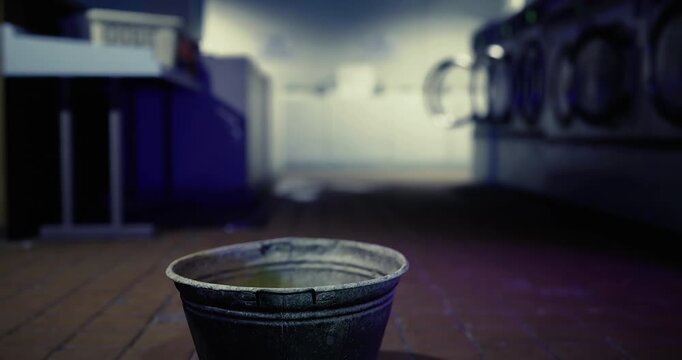 A weathered bucket rests on the floor of a dimly lit laundromat. The background shows rows of washing machines quietly humming, creating a tranquil yet eerie atmosphere.