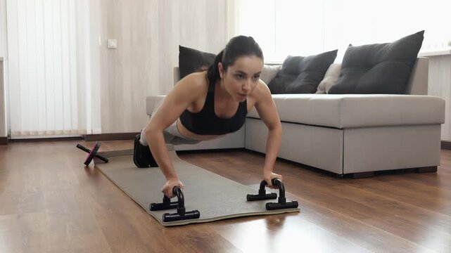 Young woman has workout at home during quarantine. Front voew of strong powerful female person doing push-ups with sports equipment. Camera moving to woman continue exercising.