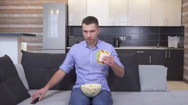Young man sit down at sofa and turn on tv. Starting to watch with interest and intensive looking. Eating sandwitch and popcorn. Chewing and looking forward.