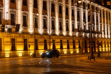 Car driving past illuminated historic building facade with classical columns and ornate architecture at night on wet city street. Urban nightscape, elegant architecture and city transportation.