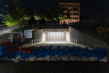 Blue and white sandbags flood barrier blocking underground parking entrance at night with illuminated gate and historic building in background. Concept of urban flood protection and emergency measures