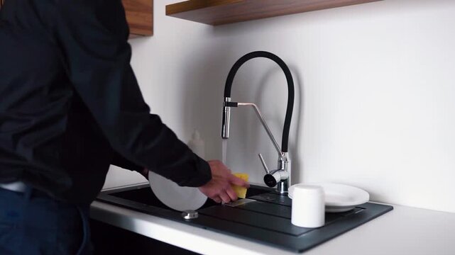 Video of man in business look open faucet and start to fash dishes or white plates with sponge. Camera moves from left side to right. Cleaning plates after lunch at home.