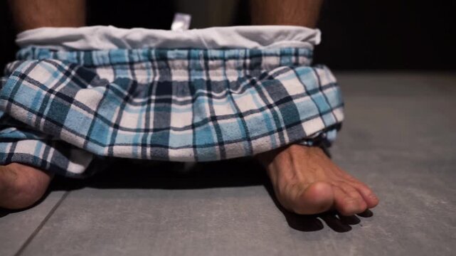 Camera moving to left. Man's feed dancing in one rythm Guy sit on pot in rest room with pants on his feet. Reaching to toilet paper roll with hand.