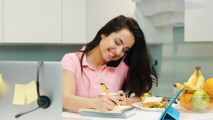 Young woman working at home during quarantine. Slow motion of multitasking girl talking on phone and making notes. Remote work office in kitchen. - Powered by Adobe