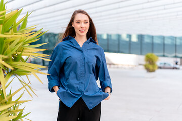 Smiling woman in blue shirt looking away at corporate workplace. Businesswoman in smart casual standing with hands in pockets, exuding confidence and professionalism