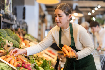 Girl salesperson adjusts carrot vegetables in stores display case. Attractive display of goods in...
