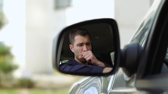 Young man inside car. A view in side mirror of guy sitting inside and caughing. Caught cold. Suffer from it in slow motion. Cover mouth with hand.