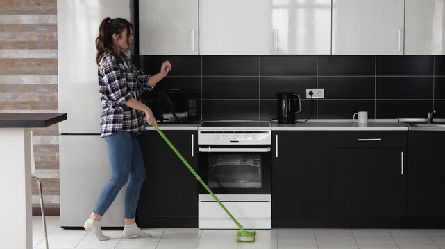 Young woman in kitchen during quarantine. Dancing and cleaning floor with mop. House wife enjoying time in kitchen.