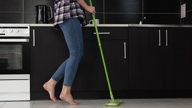 Young woman in kitchen during quarantine. Cute view motion of female legs barefeet on floor dancing back and forward. Girl hold mop and clean floor.