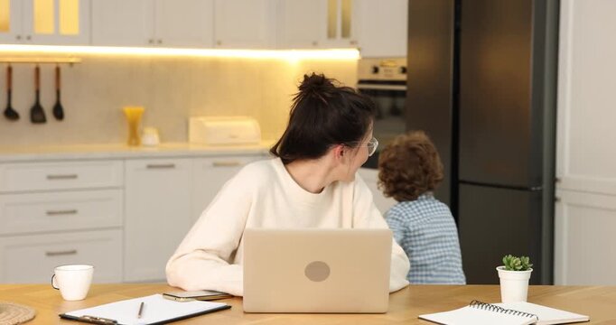 Smiling woman in glasses working with laptop while her naughty son bothering her at wooden desk in home office