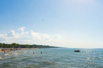 Warm sunlight bathes the beach where families and friends gather to enjoy the water. A boat glides along the calm waves as clouds dot the blue sky, creating a perfect summer atmosphere.