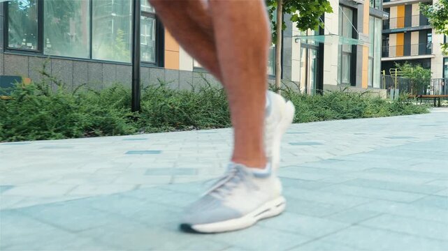 Young man exercising outside. Side view slow motion of guy's thin feet in white sneakers jogging ir slow running on street at building. Exercise training for good health and vitality.