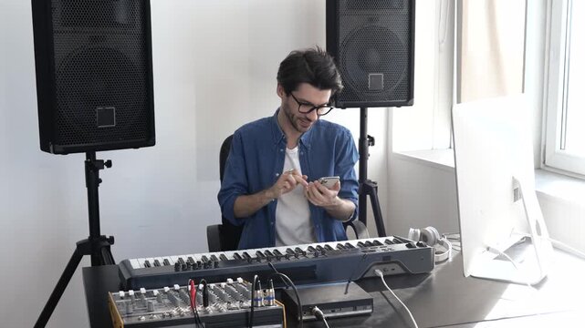 Young man in sound recording studio. Guy sit at keyboard and using smartphone. Typing on screen and smile. Prepare for playing or sound recording.