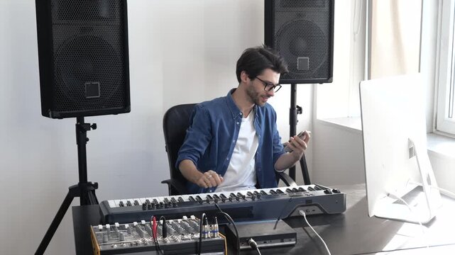 Young man in sound recording studio. Sit at keyboard and using smartphone. Guy typing on phone screen. Social media life online. Using music console or mixer.