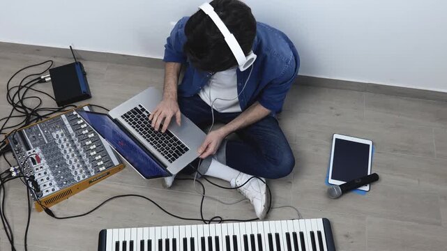 Young man in sound recording studio. Up view of guy sitting on floor and wokring with laptop. Using sound mixing console for recording. Synthesizer with microphone and tablet on floor.