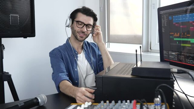 Young man in sound recording room. Guy sit on table and adjusting console in slow motion. Working with professional sound equipment. Listen to music through headphones.
