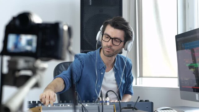 Young man in sound recording studio. Slow motion of male video blogger filming process of adjusting music mixing console and working with it. Listening to sound through headphones.