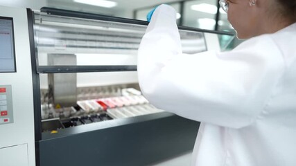 In a modern lab, a female scientist uses a tablet to monitor a large automated machine as it processes biological samples, combining human oversight with technological efficiency. - Powered by Adobe