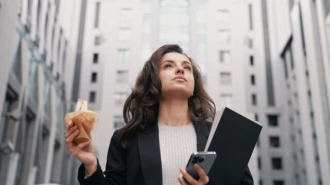 Young Caucasian woman stepping down, walking in the city eating sandwich and scrolling her smartphone. Outdoors, lunch, city center.