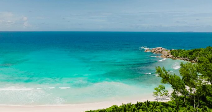 Turquoise waves gently meet a white sandy beach with distant rock formations and lush greenery. Anse Cocos. Seychelles, La Digue.