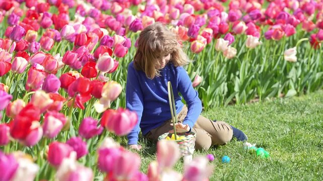Easter child in blossom spring park. Kid hunting Easter eggs near tulips. Kid play wit Ester eggs outdoor. Kids portrait in garden. Dreamy and surprised child look. Kid play with Easter eggs.