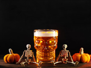 Spooky Halloween beer in a scull glass on a table with skeletons and pumpkins, copy space