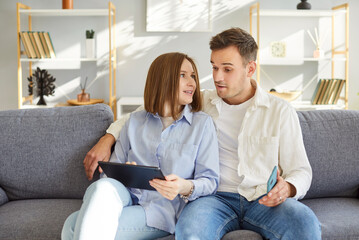 Young couple family using tablet computer together, sitting in living room, surfing internet, shopping online, making purchases, planning new room repairing, thinking about future order, choosing