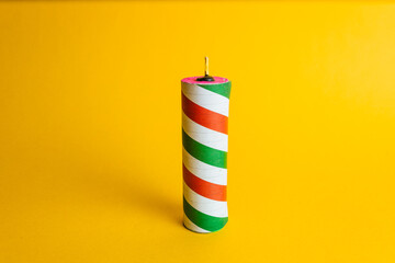 Cylindrical pyrotechnic firecracker with a red, green, and white striped design on a smooth, vibrant yellow studio background.